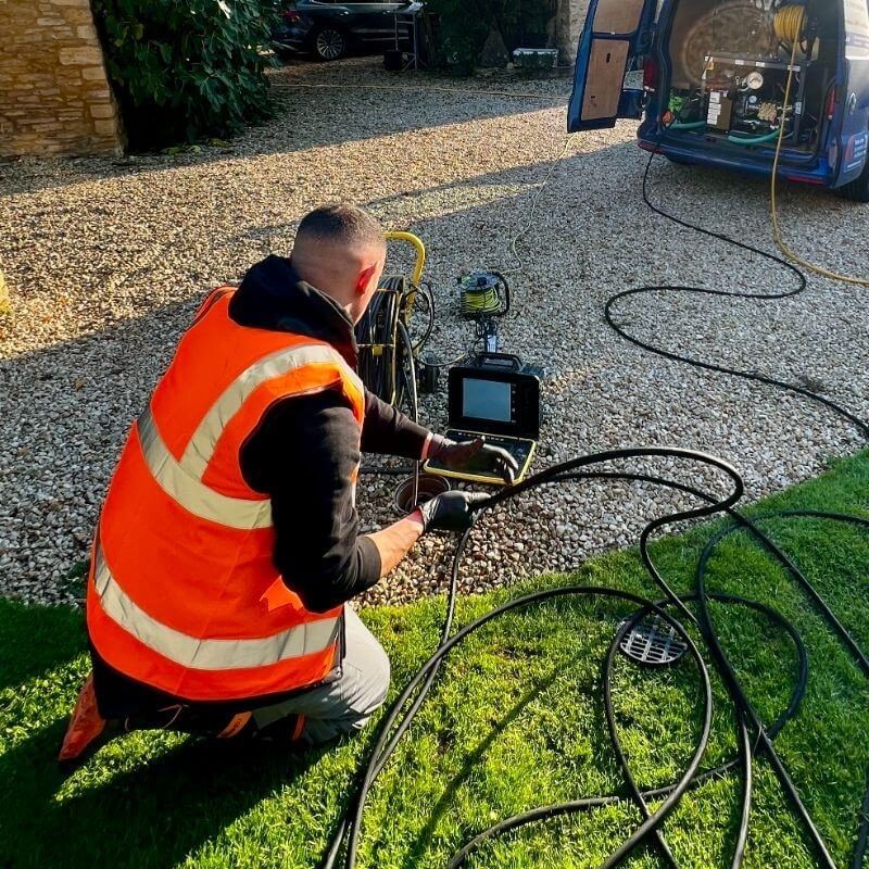 An engineer in hi-vis kneeling on a lawn with a CCTV drain camera and monitor