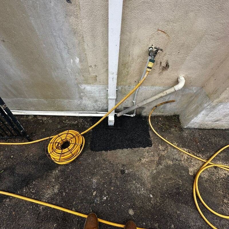 A yard gully beside a house wall with a yellow jetting hose running into it