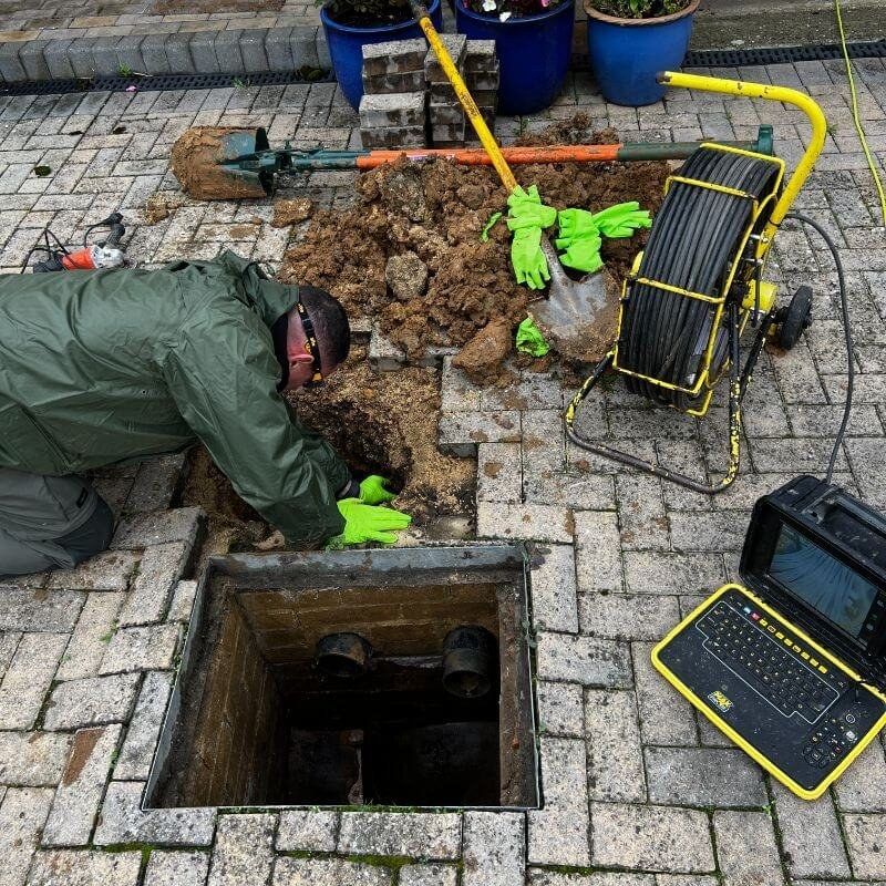 An engineer digging through block paving onto a collapsed drain pipe, with survey kit beside the hole